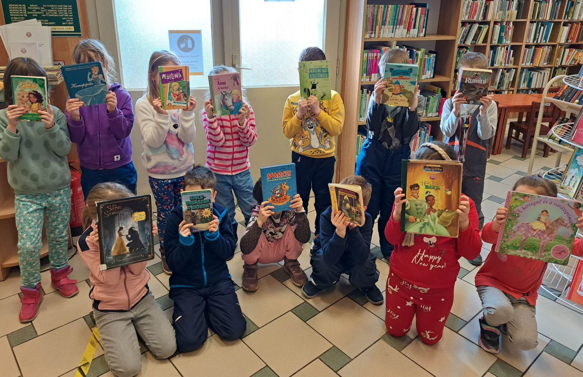 gyerekek a könyvtárban, arcuk elé könyvet tartva